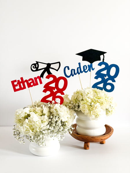 Class of 2026 personalized graduation centerpiece sticks displayed in white floral arrangements with red and blue custom name and year designs